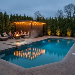 An inviting backyard scene at dusk, featuring a custom inground swimming pool, a steaming hot tub, and a covered lounge area with string lights and a fire pit where people are relaxing, emphasizing a private, year-round retreat.
